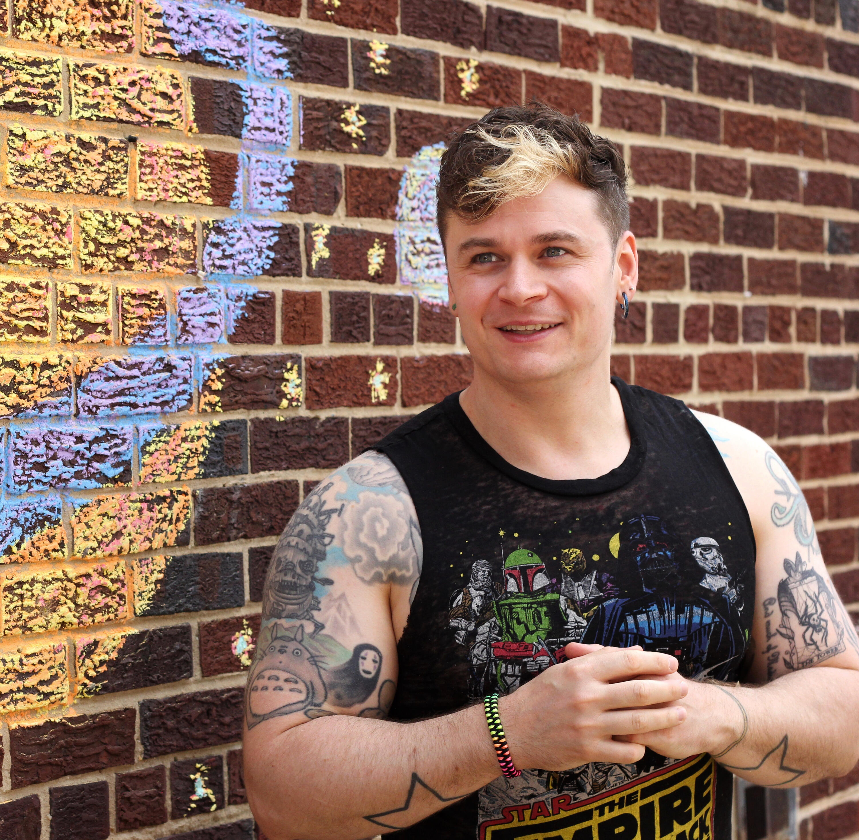 In this photograph, Trevor, wearing a sleeveless Star Wars: The Empire Strikes Back shirt, stands against a red brick wall with a chalk drawing of stars and planets on it as he stares off into the distance and smiles.