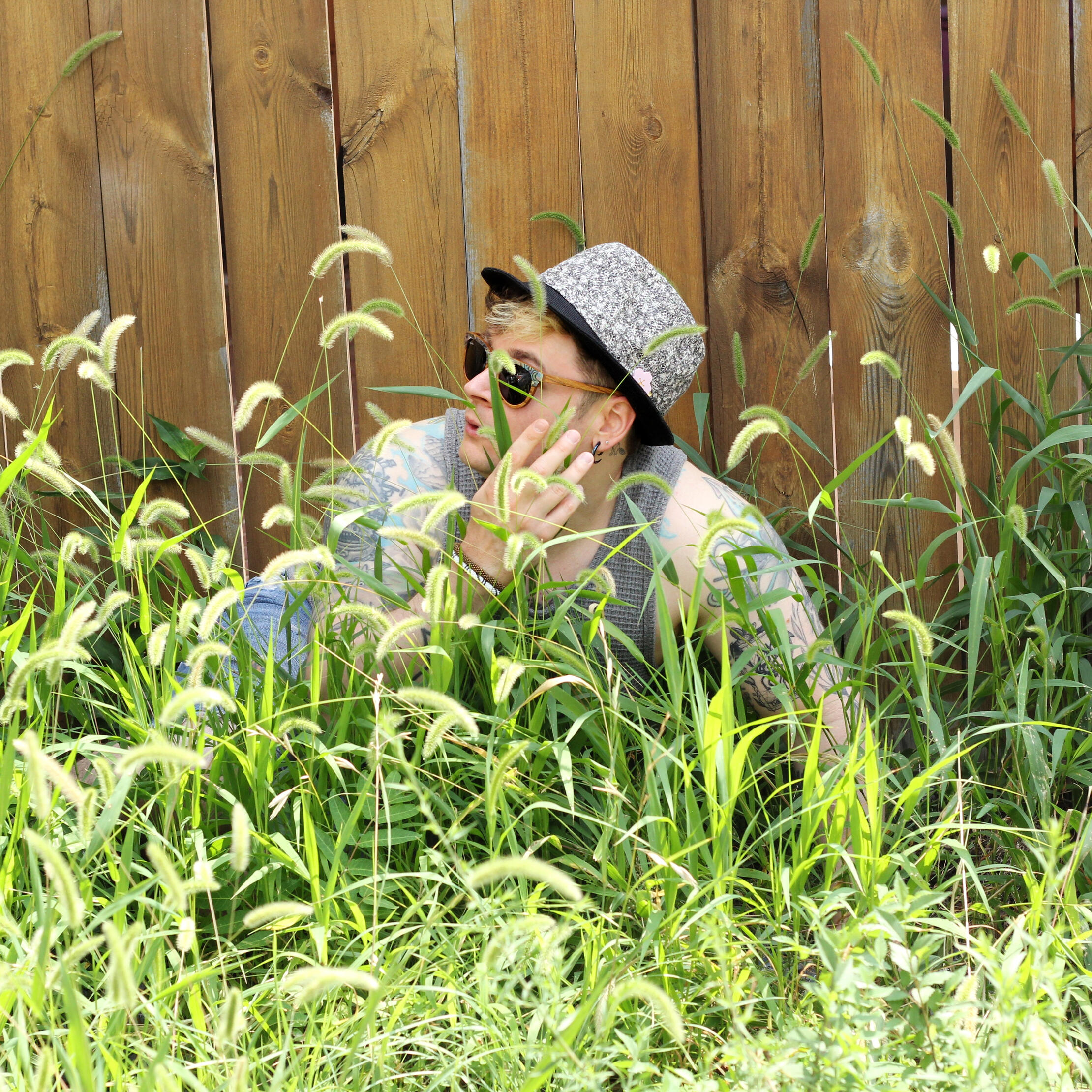 Trevor Garner, wearing some fancy wood framed sunglasses, hand made blue-grey fedora, and a waffled grey tank top, hides behind tall grass against a an old wooden fence.