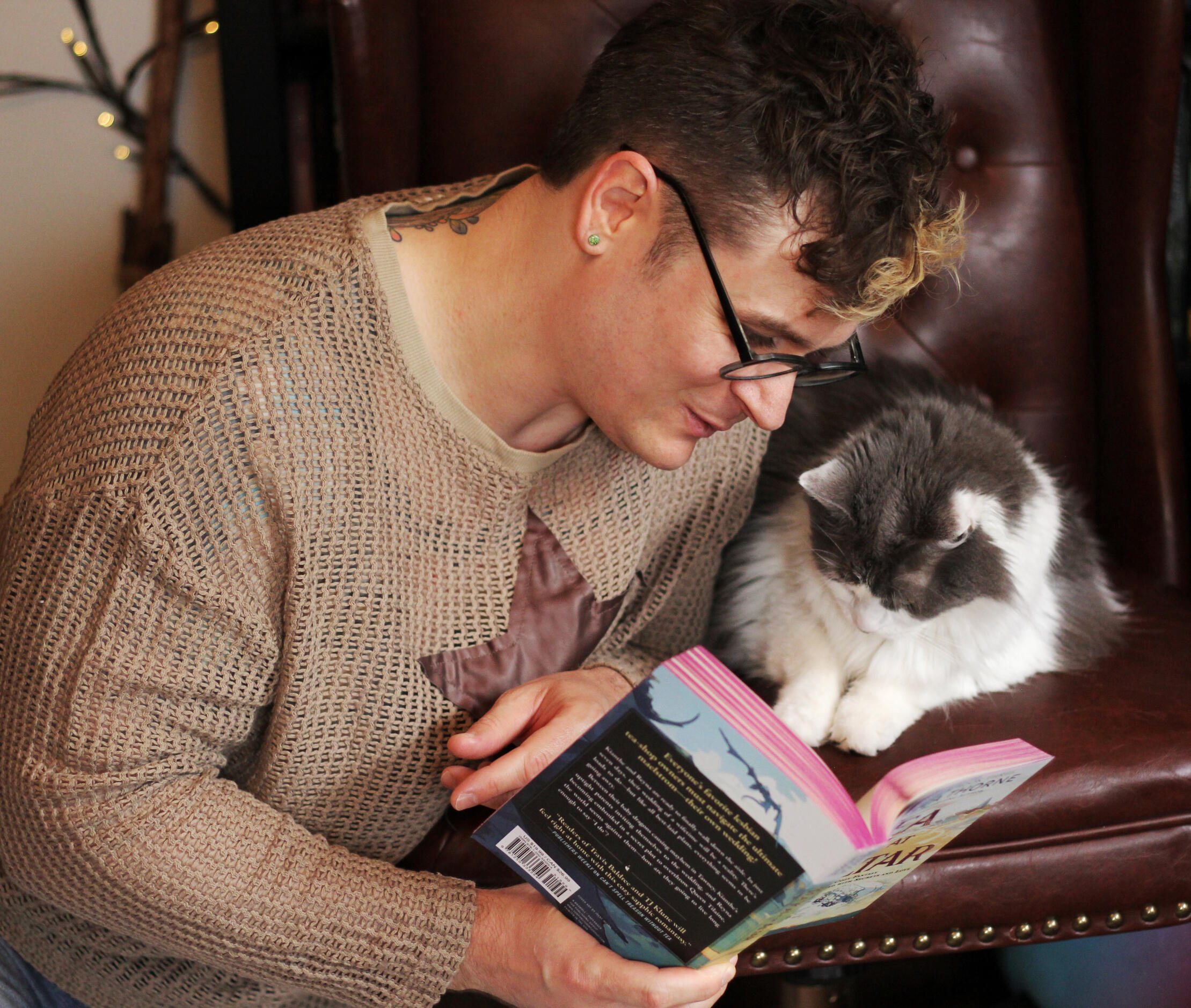 Trevor Garner, in glasses and a fishnet tan long sleeved shirt with a leather star sewn onto the front, kneels as he reads one of his favorite books to his grey and white long haired cat Sophie, who sits beside him in a chair and seems very interested in t